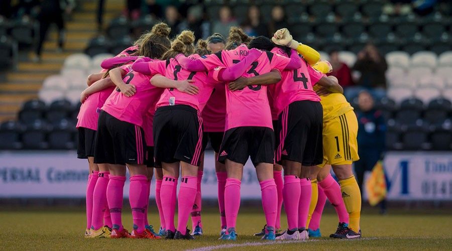 A group of female footballers in a pink and black strip, standing in a huddle.